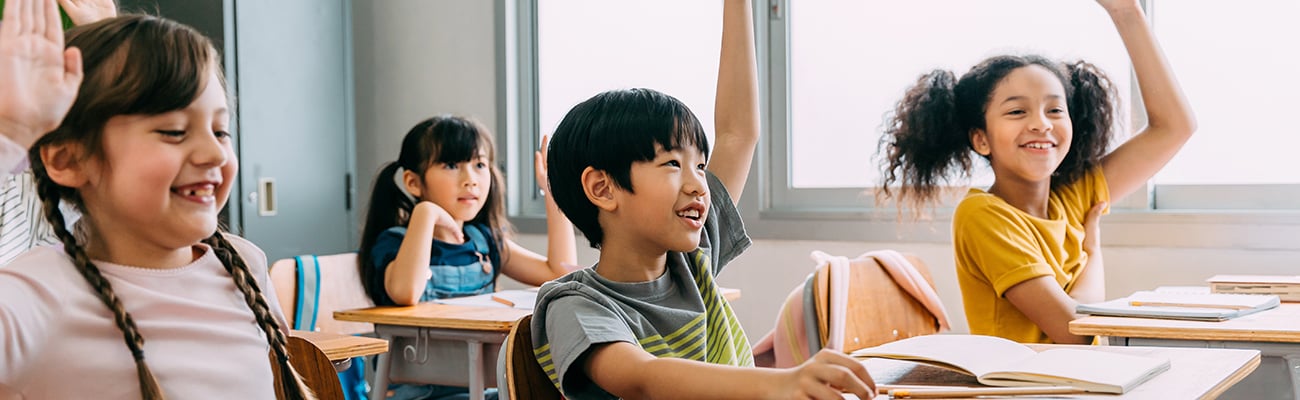 elementary students in class smiling and raising hands